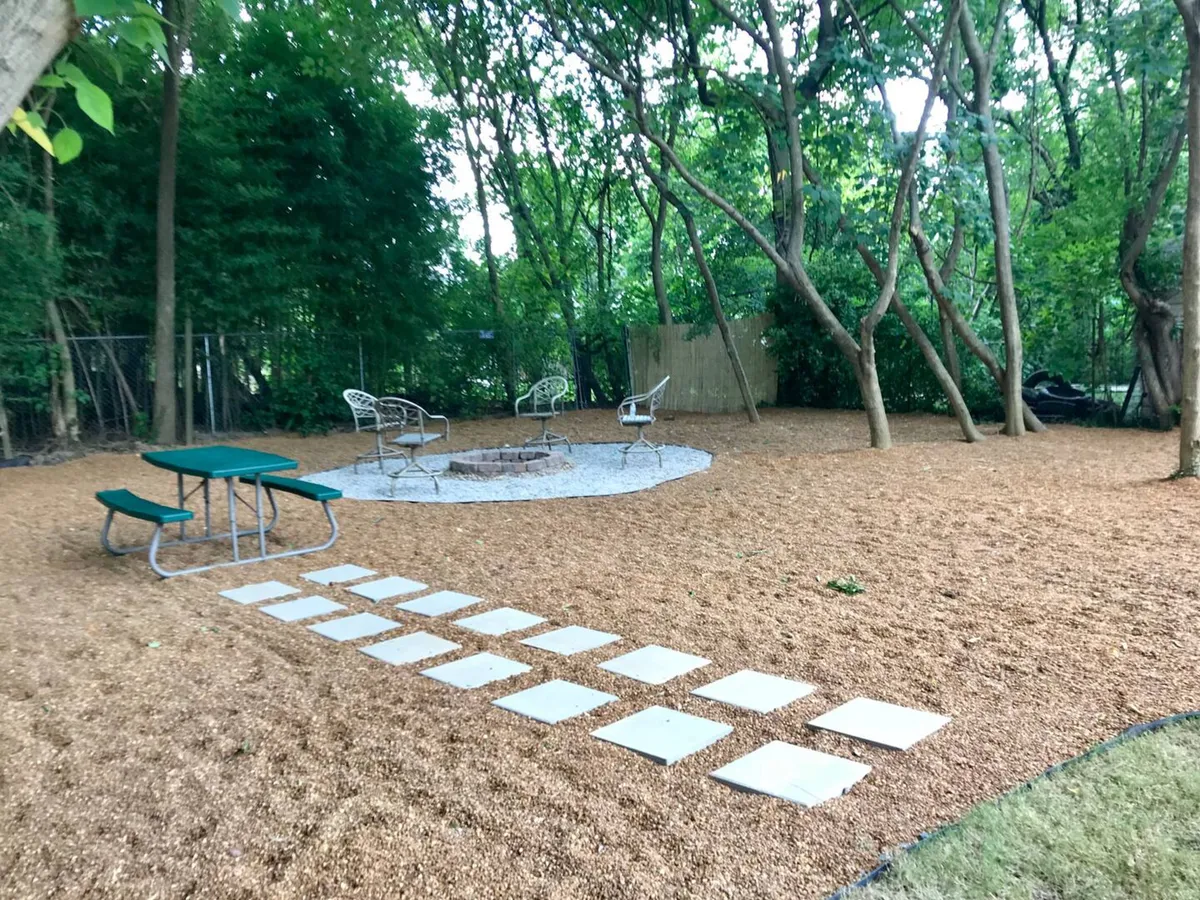 A graveled outdoor living area featuring a central fire pit surrounded by comfortable seating. The scene includes subtle details such as firewood and s’more supplies, with a relaxed, inviting mood. The area is designed for gatherings, socializing, and enjoying cozy campfire experiences in an urban setting.
