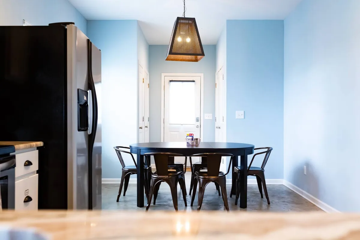 This image showcases the dining area with a prominent dining table surrounded by sleek black chairs. The background reveals modern kitchen appliances and a stylish light fixture, capturing both the functionality and inviting atmosphere of the space. The setup suggests a place for shared meals, celebrations, and engaging conversation, blending the kitchen and dining zone into one cohesive area.