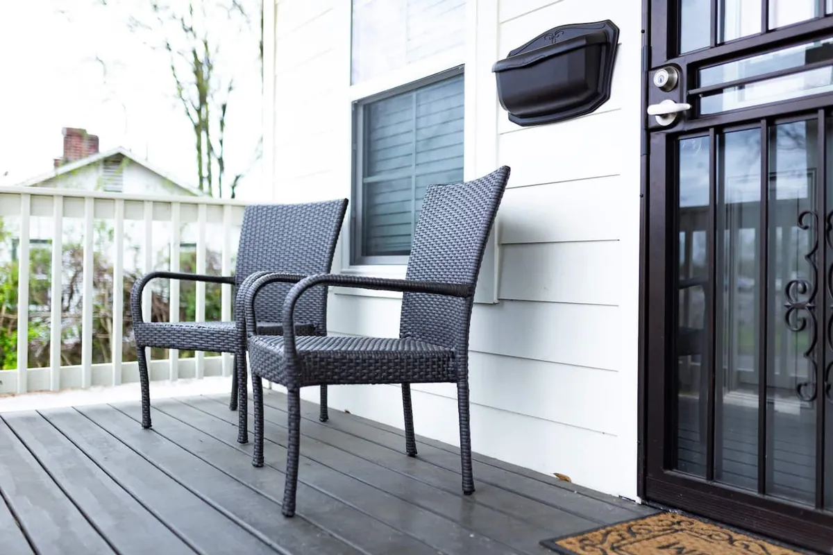 A cozy outdoor area at the house’s entrance with two wicker chairs, a welcome mat, and the front door—suggesting a peaceful spot for relaxing, socializing, or family moments just outside your door.