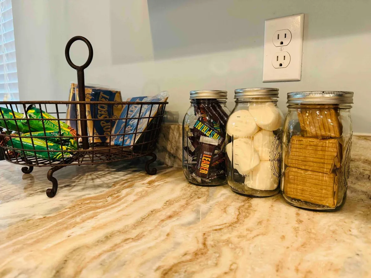 A close-up of the kitchen countertop featuring a wire basket with snacks, along with jars of marshmallows and graham crackers. The arrangement highlights the kitchen's homey and welcoming vibe, emphasizing its role as a place for snack preparation and casual gatherings.