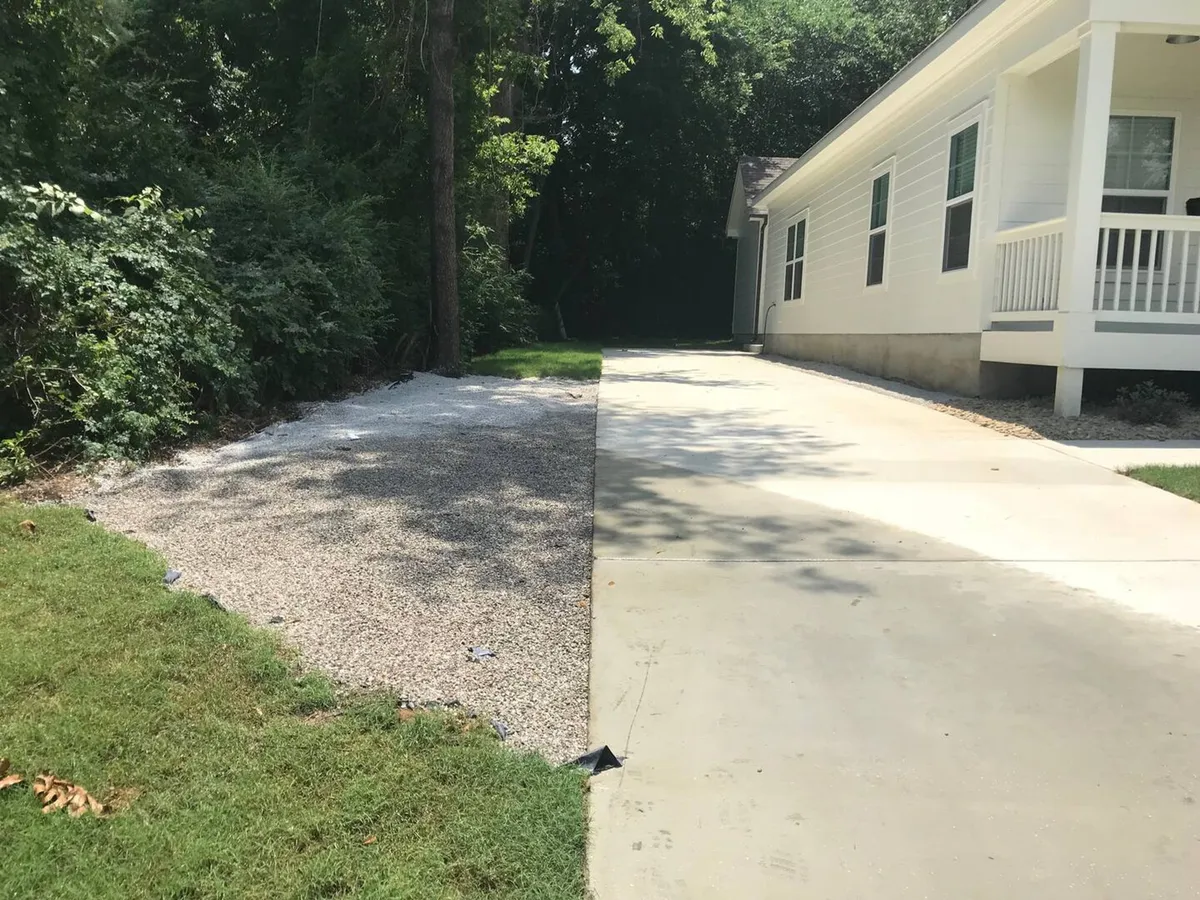 The exterior shot highlights a concrete driveway, a gravel area, and lush greenery surrounding the property. This welcoming entrance sets the stage for convenient arrivals and outdoor gatherings.