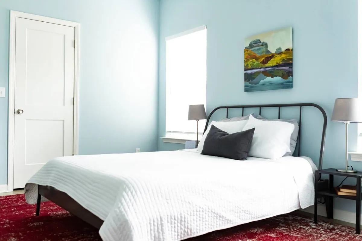A tranquil bedroom featuring a neatly made bed, a vibrant painting above the headboard, and a window letting in abundant natural light. The setting is calm and uncluttered, with soft linens and a cozy nook perfect for reading or unwinding.