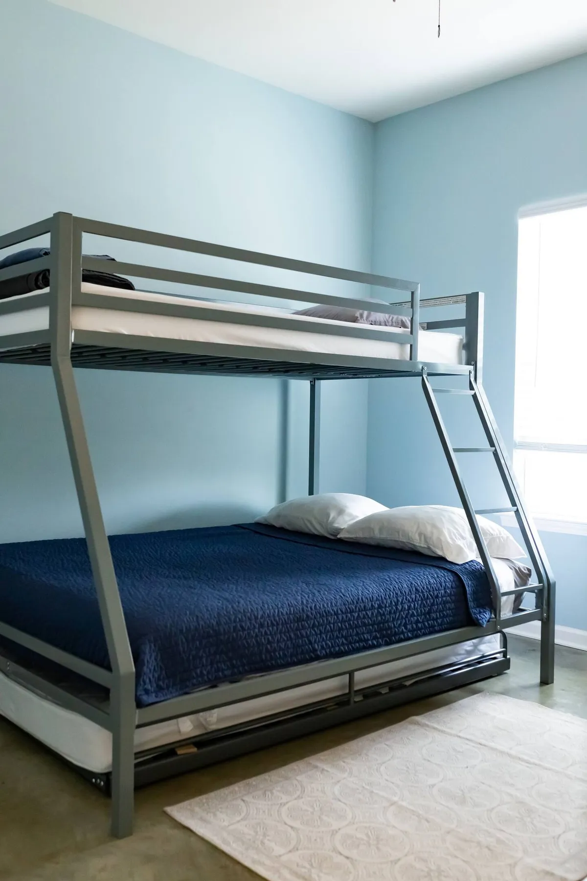 A playful, inviting bedroom with a bunk bed, blue accent walls, a window streaming in sunlight, and a colorful decorative rug. The space feels ideal for children or friends, balancing comfort and fun.