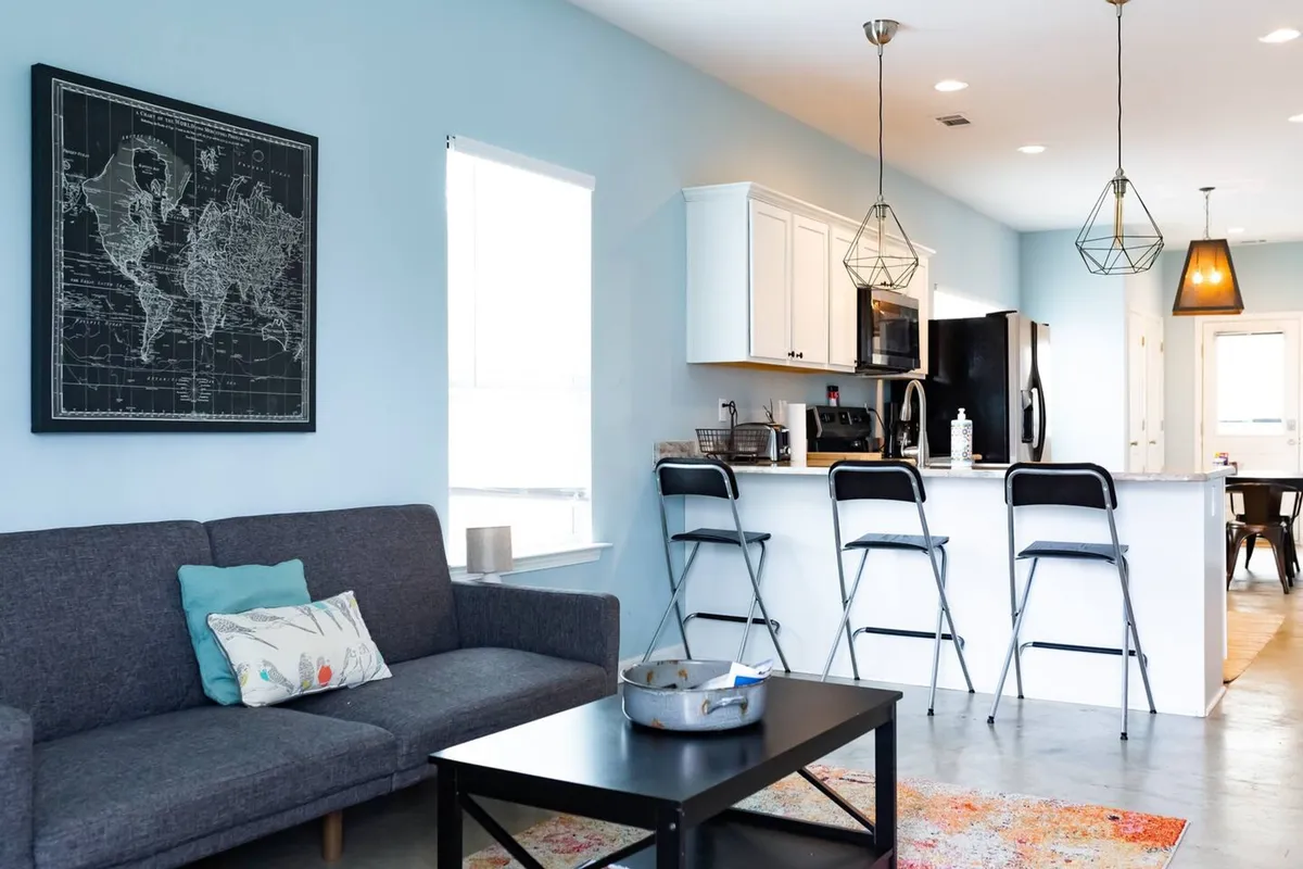 A bright, open-concept living area featuring a gray sofa arranged for conversation, a stylish world map artwork on the wall, and a kitchen island with stools visible in the background. The space includes concrete floors and large windows that invite in natural light, creating an inviting atmosphere for socializing or relaxing.